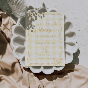 May include: A rectangular birthday invitation card featuring a yellow and white gingham pattern with a decorative border. The text reads Our Little Honey Is Turning One, Natalia's 1st Bee-Day, Saturday Aug 20, 3 o'clock, and additional details.