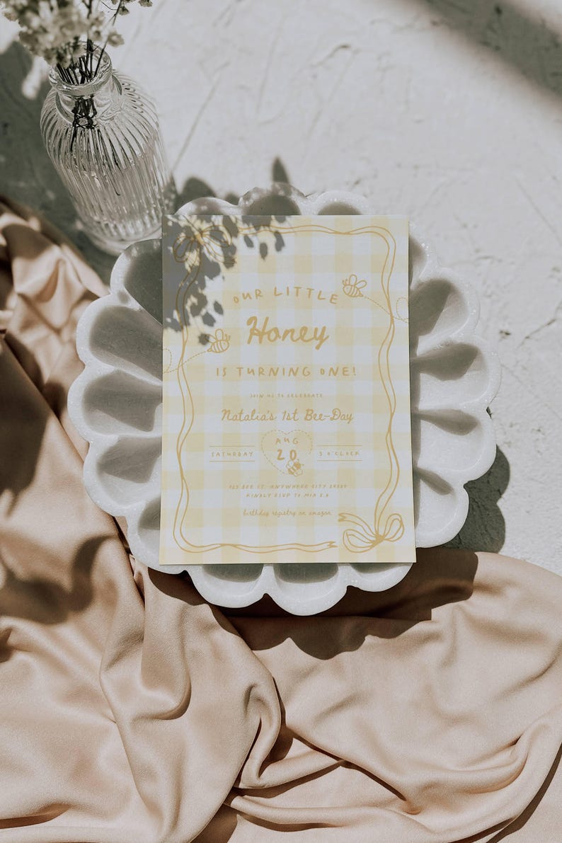 May include: A rectangular birthday invitation card featuring a yellow and white gingham pattern with a decorative border. The text reads Our Little Honey Is Turning One, Natalia's 1st Bee-Day, Saturday Aug 20, 3 o'clock, and additional details.