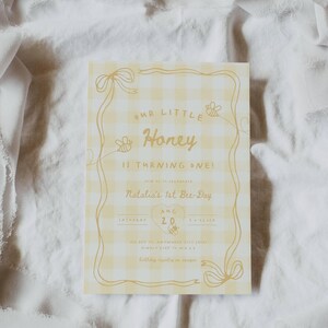 May include: A rectangular birthday invitation card featuring a yellow and white gingham pattern. The text reads Our Little Honey Is Turning One! Natalia's 1st Bee-Day, Aug 20, Saturday, 5 o'clock. It includes illustrations of bees and ribbons.