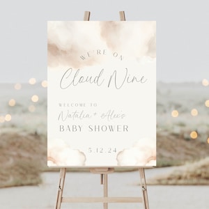 May include: A white and beige watercolour baby shower sign with the text "We're on Cloud Nine" and "Welcome to Natalia + Alexis Baby Shower 5.12.24".