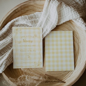 May include: Two rectangular cards with a yellow and white gingham pattern. The left card features yellow text reading Our Little Honey Is Turning One! Natalia's 1st Bee-Day, along with illustrations of bees and ribbons. The right card is blank gingham.