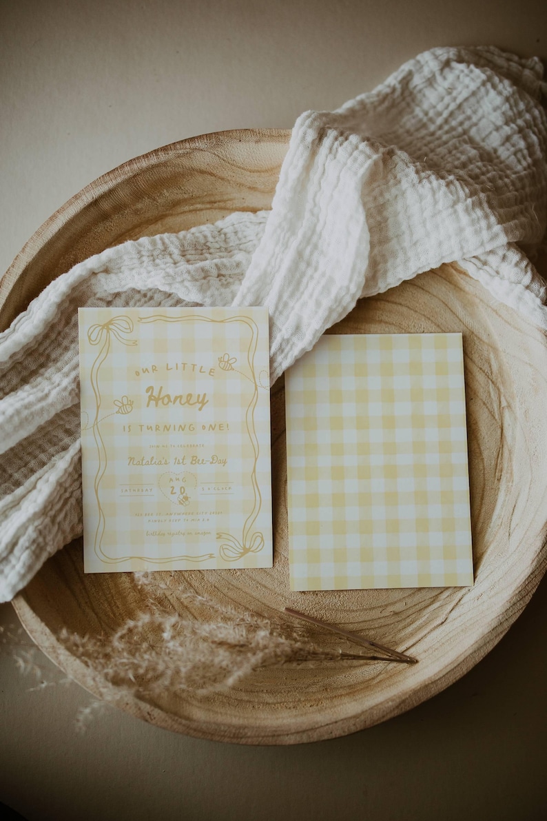 May include: Two rectangular cards with a yellow and white gingham pattern. The left card features yellow text reading Our Little Honey Is Turning One! Natalia's 1st Bee-Day, along with illustrations of bees and ribbons. The right card is blank gingham.