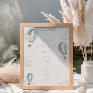 May include: A wooden framed sign for a baby shower. The sign features watercolor illustrations of blue hot air balloons and clouds. The text reads "Natalia's Baby Shower" and lists names under "Table 8".