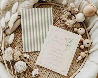 Puede incluir: Una invitación a una fiesta de bienvenida al bebé con un fondo a rayas verde y blanco. La invitación presenta una copa de martini con una aceituna y el texto "a tiny one is on the way!" y "napkin games" en una fuente cursiva. La invitación está en una cesta tejida con flores secas y juguetes de madera.