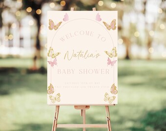 Puede incluir: Un cartel de bienvenida rosa y dorado para un baby shower. El cartel presenta un círculo con el texto "Bienvenido al baby shower de Natalia" y mariposas.