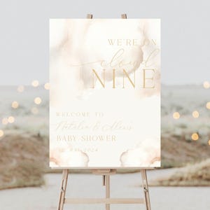 May include: A white sign with gold lettering announcing a baby shower for Natalie & Alex. The sign has a watercolour background with a cloud design and the text "We're on cloud nine". The date of the shower is listed as 12 May 2024.