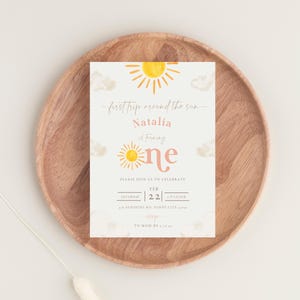 May include: A yellow and orange sun with a pink "one" on a white background with clouds. The text "first trip around the sun" and "Natalia is turning" is written in cursive. The invitation is for a first birthday party on 22nd February at 4 o'clock. The address is 456 Sunshine Rd, Sunny City 51005. RSVP by 27th April.