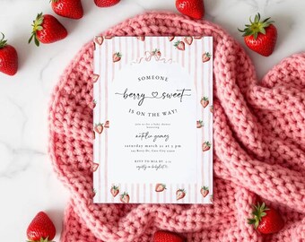 May include: A baby shower invitation with a pink and white striped design, featuring strawberries and the text "Someone Berry Sweet Is On The Way!". The invitation is placed on a pink knitted blanket, surrounded by fresh strawberries.