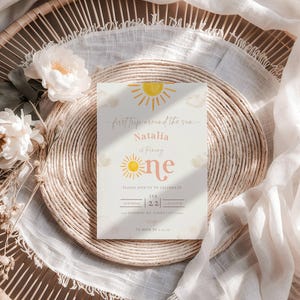 May include: A yellow and orange sun graphic with the text "first trip around the sun" and "Natalia is turning one" on a white background. The text "Please join us to celebrate" is below the sun graphic. The date and time of the event are listed below the text. The background is a light brown wicker surface.