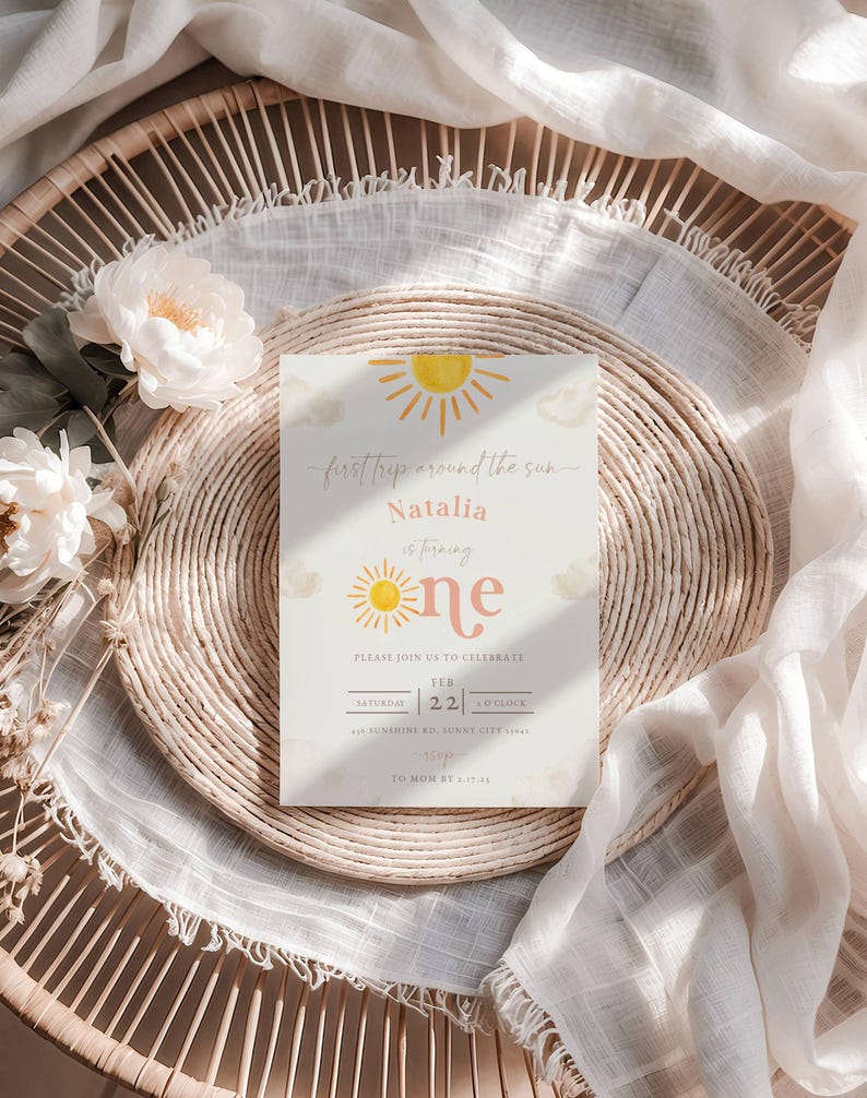 May include: A yellow and orange sun graphic with the text "first trip around the sun" and "Natalia is turning one" on a white background. The text "Please join us to celebrate" is below the sun graphic. The date and time of the event are listed below the text. The background is a light brown wicker surface.