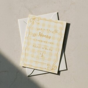 May include: A rectangular invitation card with a light yellow gingham pattern and gold text. The card features a decorative gold border with ribbon accents and small bee illustrations. The text reads Our Little Honey Is Turning One, Natalia's 1st Bre-Day, and includes date details. An envelope is placed underneath.