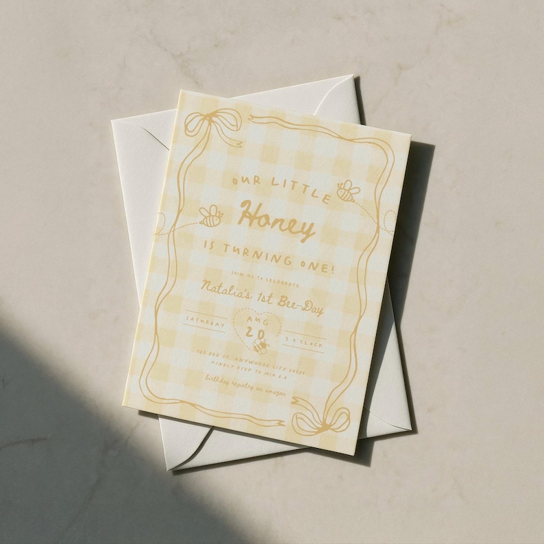 May include: A rectangular invitation card with a light yellow gingham pattern and gold text. The card features a decorative gold border with ribbon accents and small bee illustrations. The text reads Our Little Honey Is Turning One, Natalia's 1st Bre-Day, and includes date details. An envelope is placed underneath.