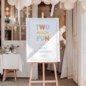 May include: A rectangular white sign with a light blue scalloped border on a wooden easel. The sign features the text "TWO MUCH FUN" in colorful bubble letters, followed by "WELCOME TO ALEX'S 1ST BIRTHDAY!" and the date "7.2.20XX" in blue and orange.