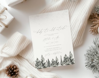 May include: A baby shower invitation with the text "baby it's cold outside" in elegant script. The invitation features a winter forest illustration at the bottom and includes event details. The invitation is surrounded by white gifts, a white sweater, and pine accents.
