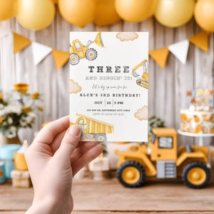 Peut inclure: Une invitation d'anniversaire sur le thème de la construction, tenue dans une main. L'invitation indique "THREE AND DIGGIN' IT!" avec les détails du 3e anniversaire d'Alex. L'arrière-plan comprend des ballons jaunes, un gâteau et un camion de chantier jouet.