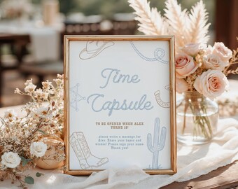 May include: A framed sign with a gold border, featuring the words "Time Capsule" in a script font. The sign includes illustrations of a cowboy hat, rope, horseshoe, boot, and cactus. Text below reads "To be opened when Alex turns 18!"