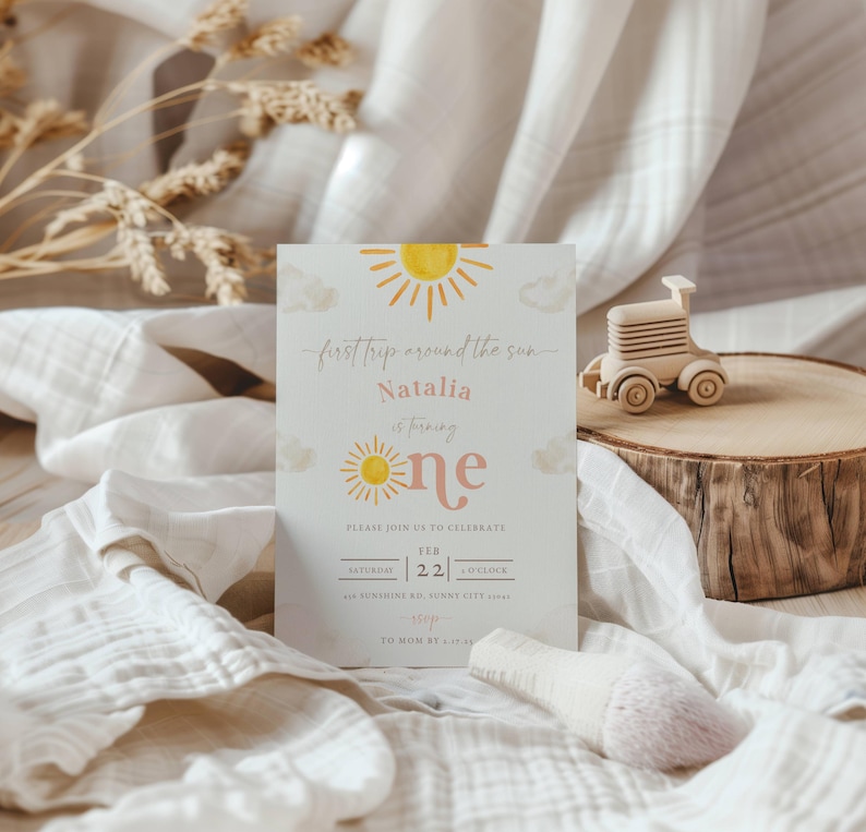 May include: A light blue invitation card with a yellow sun and clouds. The text reads "first trip around the sun Natalia is turning one" and includes the date and time of the event.