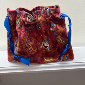 May include: A burgundy drawstring bag with a paisley pattern in shades of gold, blue, and white. The bag has bright blue ribbon drawstrings tied into bows. The bag is sitting on a white surface.