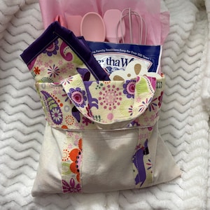 May include: A beige fabric tote bag with floral patterned accents, holding pink kitchen utensils, a package of baking mix, and a floral patterned napkin. The bag is lined with pink tissue paper.