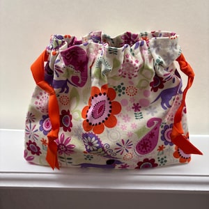 May include: A drawstring bag with a floral pattern in shades of pink, purple, orange, and green on a cream background. The bag is gathered at the top with bright orange ribbon ties.