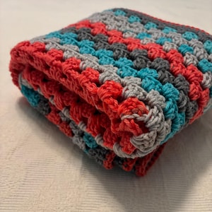May include: Folded crochet blanket in a granny square pattern. The blanket features a color scheme of red, teal, gray, and charcoal. The blanket is folded neatly, showcasing the texture and stitch detail.