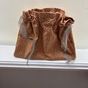 May include: A small, drawstring bag made of tan fabric with a repeating pattern of small red flowers. The bag has a gathered top and is tied with gray ribbons. The bag is sitting on a white surface.