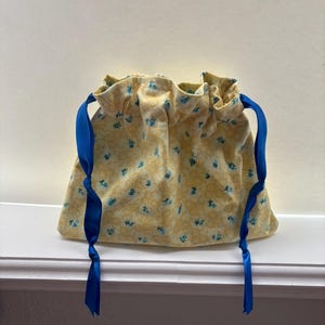 May include: A yellow drawstring bag with a floral pattern in blue. The bag is made of fabric and has bright blue ribbon drawstrings. The bag is sitting on a white ledge.