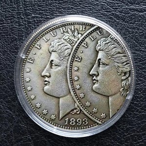 Może przedstawiać: Srebrna moneta z dwoma profilami zwróconymi ku sobie, umieszczona w przezroczystym plastikowym uchwycie. Moneta zawiera napis "E PLURIBUS UNUM" i rok "1893". Projekt monety obejmuje gwiazdy na obrzeżu.