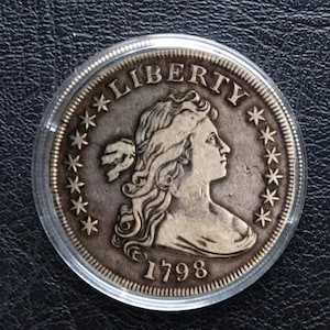 Op de afbeelding: Een zilveren munt met het woord "LIBERTY" bovenaan en het jaartal "1798" onderaan. De munt toont een profiel van een vrouwenhoofd omringd door sterren. De munt is ingekapseld in een doorzichtige plastic houder.