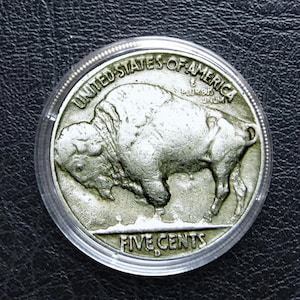 May include: A silver United States Buffalo nickel coin, encased in a clear protective holder. The coin features a detailed profile of a bison and the words "UNITED STATES OF AMERICA" and "FIVE CENTS".