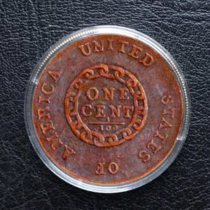 May include: A copper-colored United States one-cent coin, encased in a clear protective holder. The coin features the words "UNITED STATES OF AMERICA" and "ONE CENT" in a circular design.