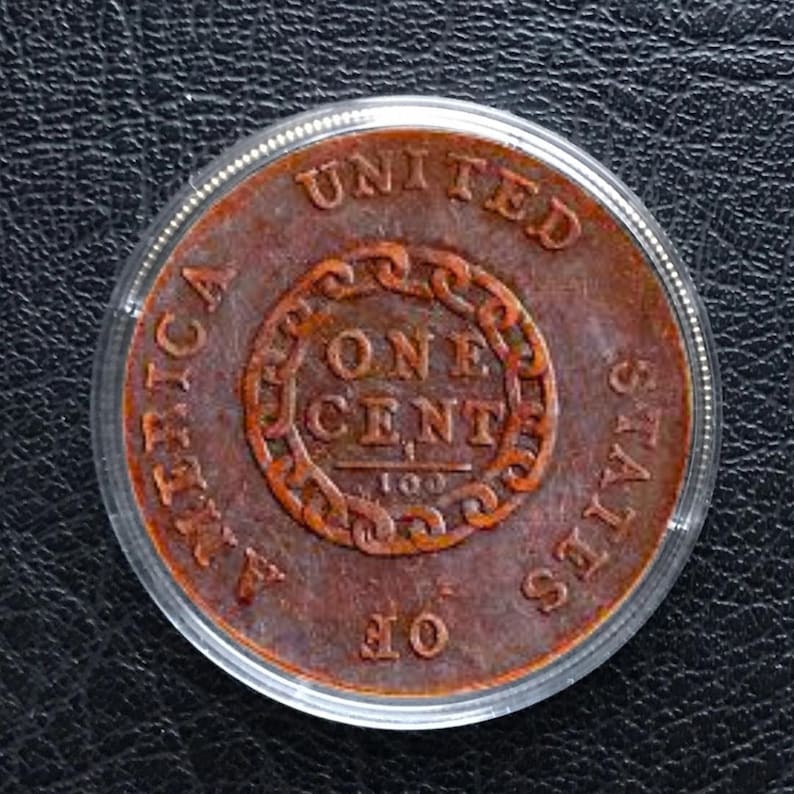 May include: A copper-colored United States one-cent coin, encased in a clear protective holder. The coin features the words "UNITED STATES OF AMERICA" and "ONE CENT" in a circular design.