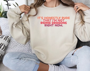 May include: A cream-colored sweatshirt with the text "It's honestly rude that I'm not riding dragons right now." in a gradient of pink and red.