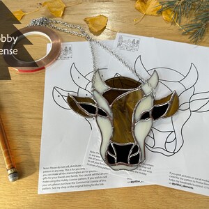 May include: A stained glass pattern of a cow's head with brown, white, and black glass pieces. The pattern is printed on white paper with instructions for making the stained glass art. The image also includes a roll of copper foil tape, a glass cutter, and a silver chain.