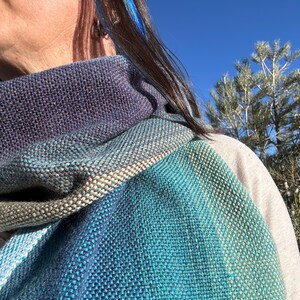 May include: A woven scarf in shades of blue, teal, and gray. The scarf is wrapped around the neck, showcasing a textured pattern. The colors blend together, creating a gradient effect. The scarf appears soft and comfortable.