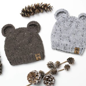 Può includere: Due berretti di lana con orecchie da orso. Un berretto è grigio con macchie nere e l'altro è marrone con macchie nere. Entrambi i berretti hanno una piccola etichetta marrone con un logo ad albero.