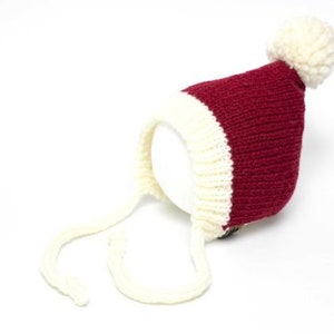 May include: A red and white knitted baby hat with a white pom pom on top and white ties. The hat is designed to look like a Santa hat.