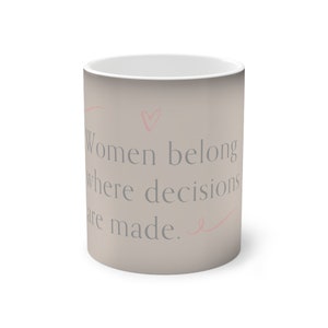 Könnte beinhalten: Eine beige Keramiktasse mit einer grauen Aufschrift, die lautet "Frauen gehören dorthin, wo Entscheidungen getroffen werden." Die Aufschrift ist von rosa Herzen und Schnörkeln umgeben.