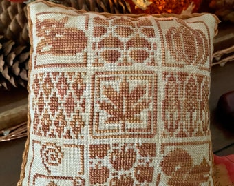 May include: A small, square, beige cushion with a brown embroidered design of autumn leaves, acorns, and pumpkins. The cushion is surrounded by autumn foliage.