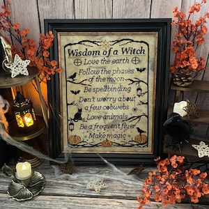 Könnte beinhalten: Gerahmte Kreuzstichkunst mit dem Text "Wisdom of a Witch" und inspirierenden Phrasen. Das Kunstwerk zeigt schwarze Stiche auf beigem Hintergrund, gerahmt in Schwarz. Dekorative Elemente sind Kürbisse, Fledermäuse und eine schwarze Katze.