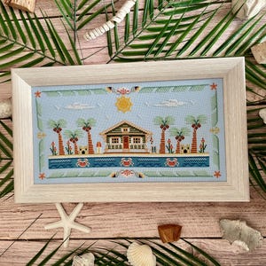 Puede incluir: Un punto de cruz enmarcado que representa una escena de playa con una casa, palmeras y cangrejos. La obra de arte presenta un fondo azul claro con un sol amarillo y nubes blancas. El marco es de color madera clara.