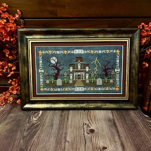 May include: A framed cross-stitch embroidery featuring a haunted house scene on a dark blue background. The design includes a mansion, tombstones with R.I.P. text, a full moon, lightning, bare trees, and a border of pumpkins and skulls.