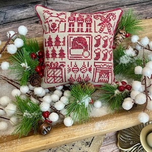 Peut inclure: Un petit coussin carré sur le thème de Noël avec un motif de point de croix rouge et beige. Le motif comprend des images du bonnet du Père Noël, des sapins de Noël et des bas. Le coussin est entouré de fausse verdure, de baies blanches et de pommes de pin.