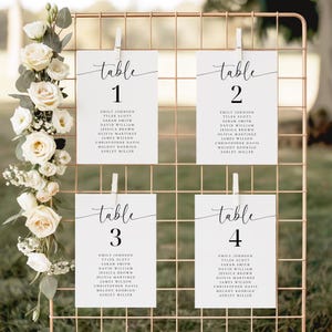 May include: A wedding seating chart with four white cards clipped to a gold wire grid. Each card is labeled "table" with a number and guest names. White roses and greenery decorate the side.