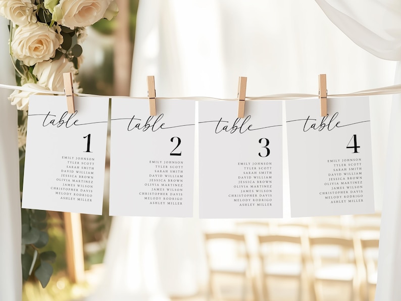 May include: Four white cards with black text are clipped to a string. Each card reads "table" in cursive, followed by a number 1-4. Below the number are names. The cards are part of a wedding or event table setting.