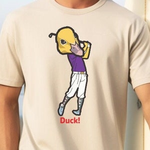 Peut inclure: Un t-shirt beige avec une image de dessin animé d'un canard portant une chemise violette et balançant un club de golf. Le texte "Duck!" est imprimé sous l'image.