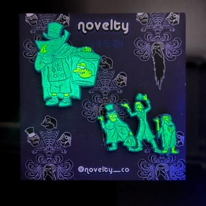 May include: Two glow-in-the-dark enamel pins featuring characters in top hats and capes, one holding a bucket with a face, displayed on a dark purple backing card with the text novelty, glow-in-the-dark, and @novelty_co printed in white.