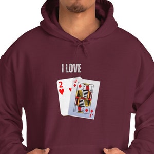 Puede incluir: Una sudadera con capucha color burdeos con un gráfico blanco de dos cartas de juego. El texto "I LOVE" está impreso sobre las cartas.