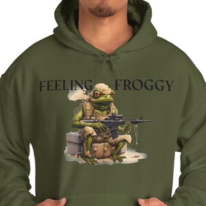 Puede incluir: Sudadera con capucha verde oliva con la frase "FEELING FROGGY" estampada. El gráfico muestra una rana con uniforme militar sosteniendo un rifle. La sudadera tiene un bolsillo delantero y una capucha con cordón.