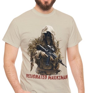 Könnte beinhalten: Ein beiges T-Shirt mit einem Soldaten in Tarnkleidung, der ein Gewehr hält. Der Soldat trägt einen Tarnanzug und hat ein Zielfernrohr auf dem Gewehr. Der Text "DESIGNATED MARKSMAN" ist auf dem T-Shirt aufgedruckt.